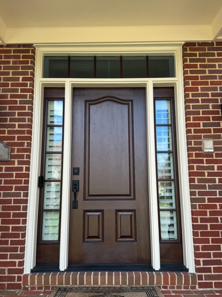Mahogany wood front entry door with arched top panel and shuttered sidelights on a brick home.